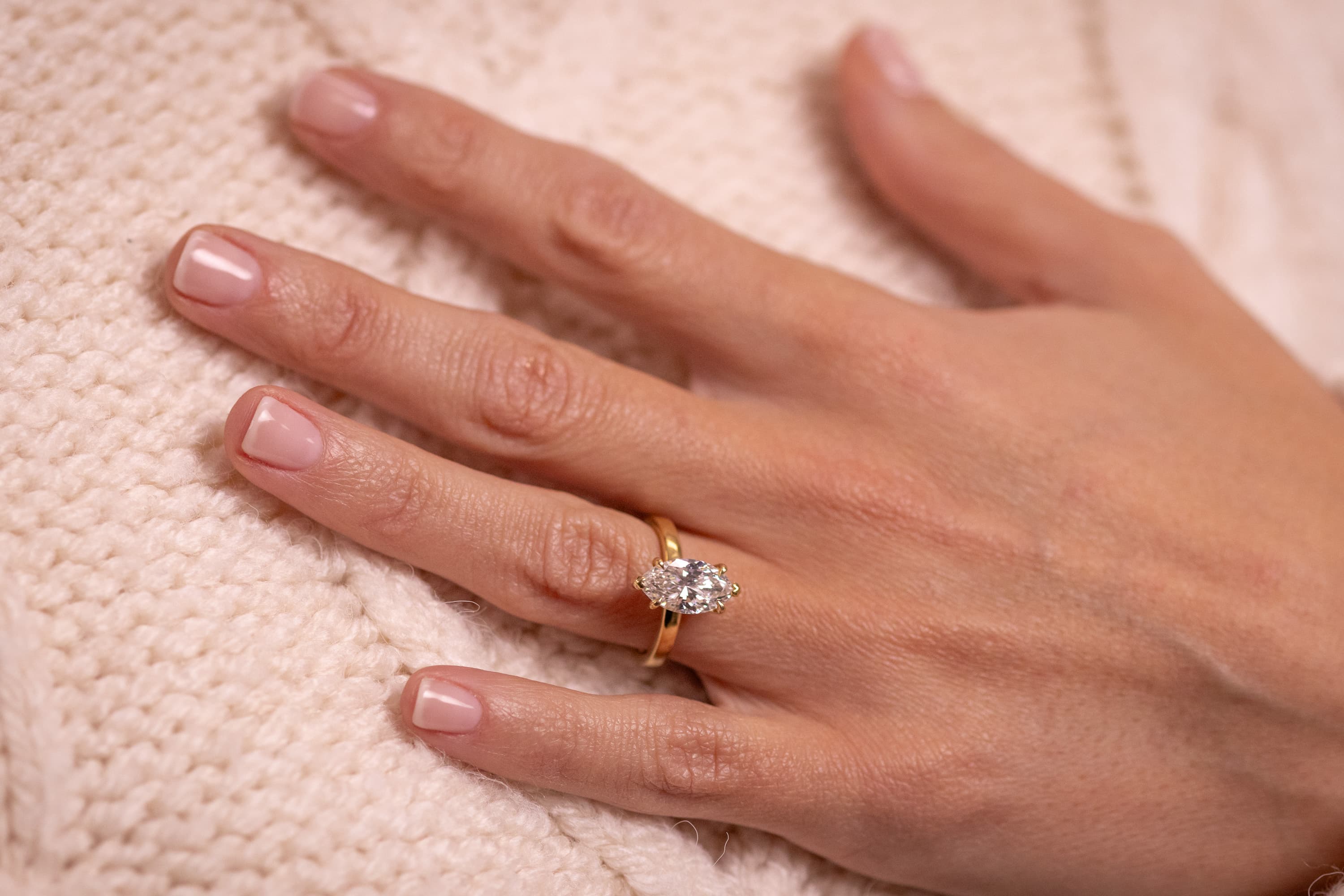 An elongated moval-cut diamond solitaire in a yellow gold band, worn on a hand resting on cream knit fabric